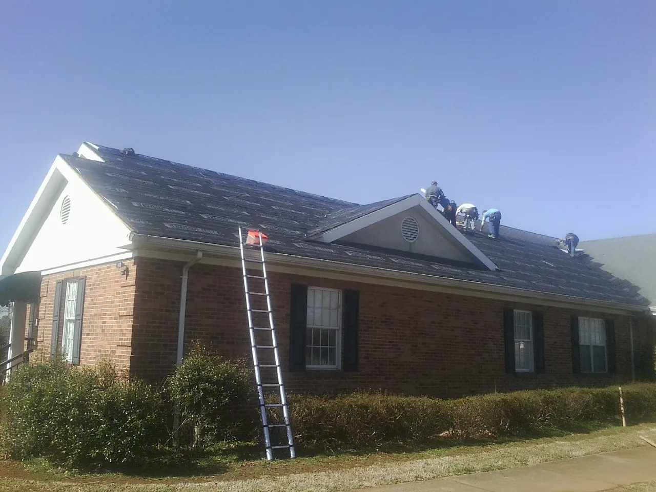 Crew tearing shingles off a brick ranch roof with a ladder set — Emergency Roof Repair work by our crew in Post
